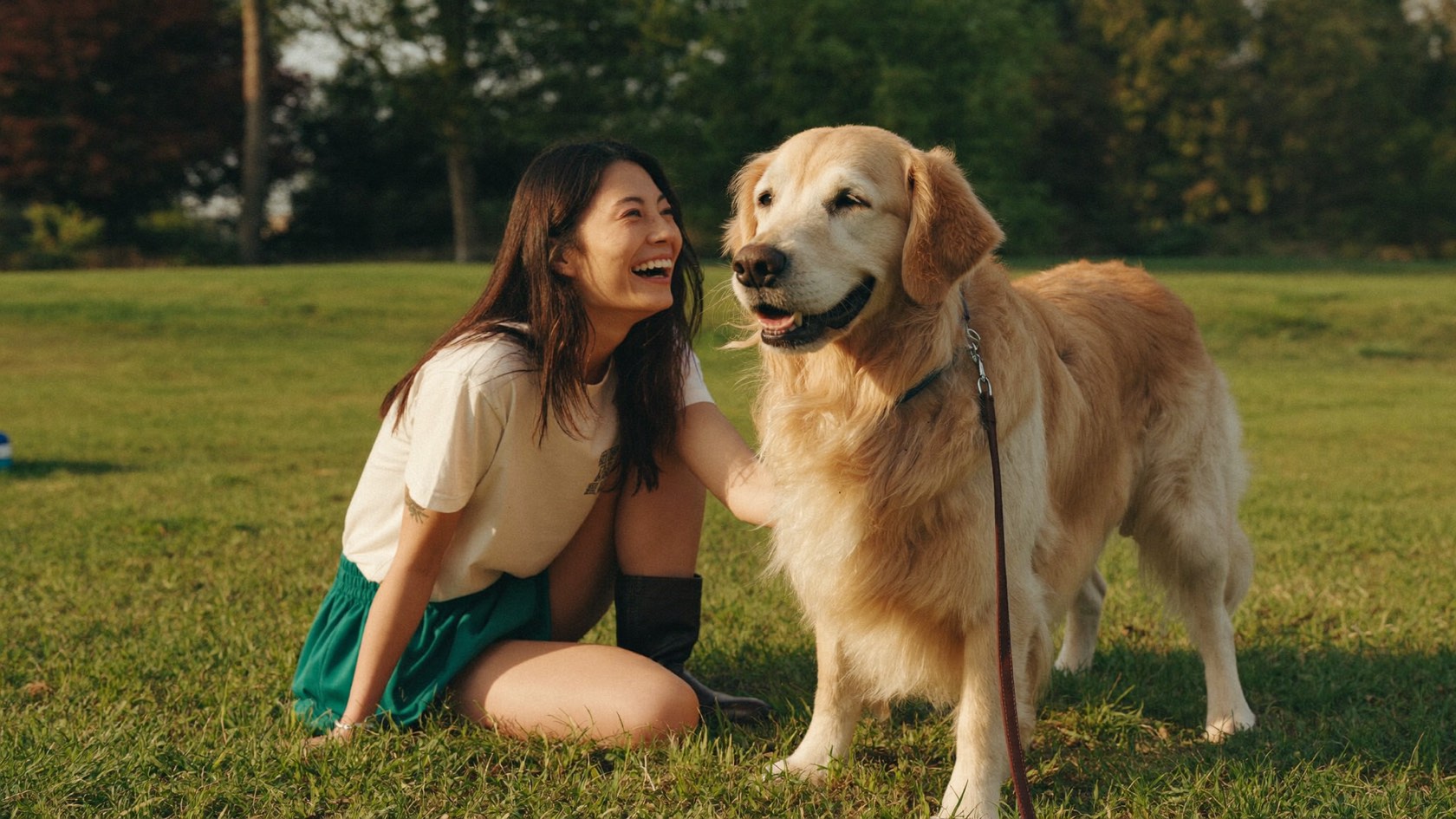 A person laughing beside her golden retriever in a sunlit park